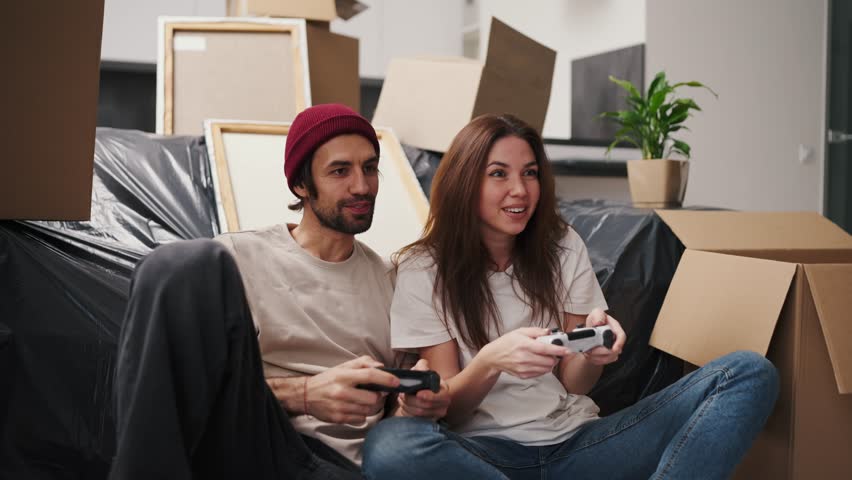 A happy and engaged couple, a brunette girl and her boyfriend with stubble in a beige T-shirt play video games using two joysticks while sitting near a sofa covered with a plastic cover and among a Royalty-Free Stock Footage #3460223277