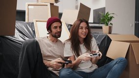 A happy and engaged couple, a brunette girl and her boyfriend with stubble in a beige T-shirt play video games using two joysticks while sitting near a sofa covered with a plastic cover and among a