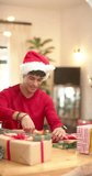 Vertical video of biracial man wrapping christmas present in decorative paper at home, slow motion. Lifestyle, christmas, celebration, gift and domestic life, unaltered.