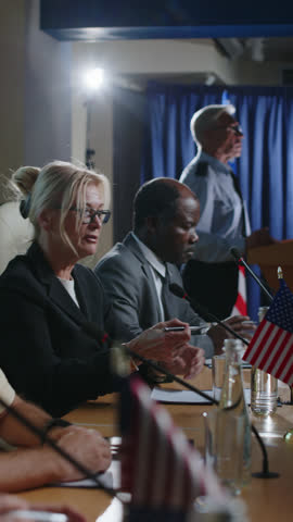 Vertical shot of senior female politician representing USA sitting at table by mic and talking during press conference
