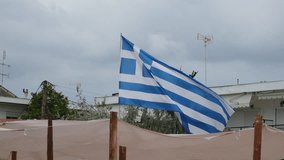 Flag of Greece flutters in wind over beach canopy against cloudy sky. Greek resort town with low houses and trees. - Powered by Shutterstock - Get 15% off with code: PIKWIZARD15