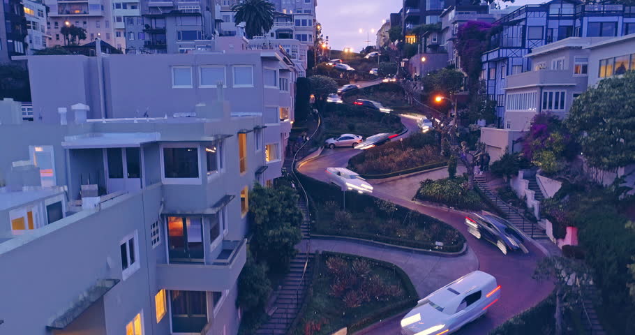 Aerial drone boom down of cars driving down the winding road of Lombard Street at night with streaky car lights. In the background is the San Francisco Skyline with a view to the Bay Bridge