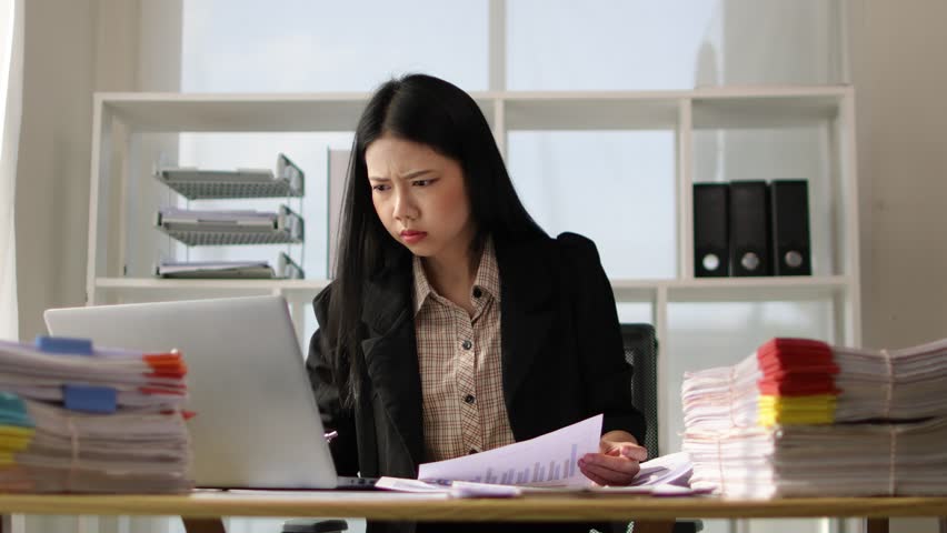 Serious Asian businesswoman is busy with paperwork on a messy desk. Woman are stressed due to excessive workload and are forced to overworked. Royalty-Free Stock Footage #3460522581