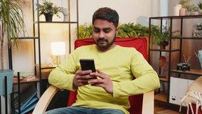 Indian man sits on chair uses mobile phone smiles at home room apartment. Young Arabian guy texting share messages content on smartphone social media applications online watching relax movie video