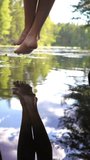 A vertically-oriented video. Young woman enjoying silence by the forest lake sitting on the edge of a wooden jetty, swinging one's feet near the water surface. A vertically-oriented video.