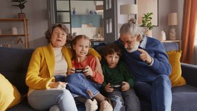 Caring grandparents sitting on sofa with happy children using console for having fun together at living room. Stylish old couple babysitting kids and gesturing with hands while watching gameplay.
