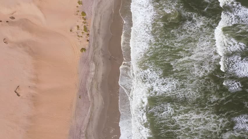 Top down aerial drone cinematic film of crashing waves from ocean onto golden sand beach shore. white foam and spray, red sand dunes in Skeleton Coast, Namibia Royalty-Free Stock Footage #3460693075