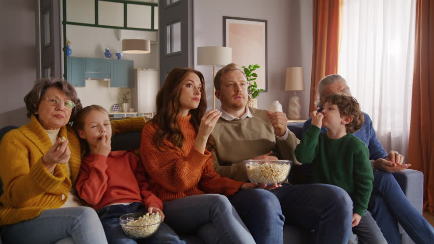Friendly family sitting on couch, eating popcorn from glass bowls and pointing with finger to tv screen. Three generations of people gathering in living room and spending leisure together in calm.