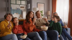 Friendly family sitting on couch, eating popcorn from glass bowls and pointing with finger to tv screen. Three generations of people gathering in living room and spending leisure together in calm. - Powered by Shutterstock - Get 15% off with code: PIKWIZARD15