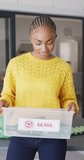 Vertical video of happy african american woman with glass recycling box. Lifestyle, spending free time at home concept.