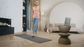 Sporty blond hair caucasian woman sits on yoga mat in living room, female fitness coach doing asana teaching yoga online, laptop video call, remote training at home, dolly wide angle shot