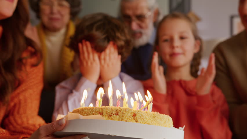 Little kid with curly hair covering face with palms and blowing all burning candles with help of older sister. Friendly family cheering up for birthday boy while looking at freshly baked cake.