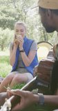 Vertical video of happy diverse couple camping, drinking tea and playing guitar in park, slow motion. Nature, hiking and park concept.