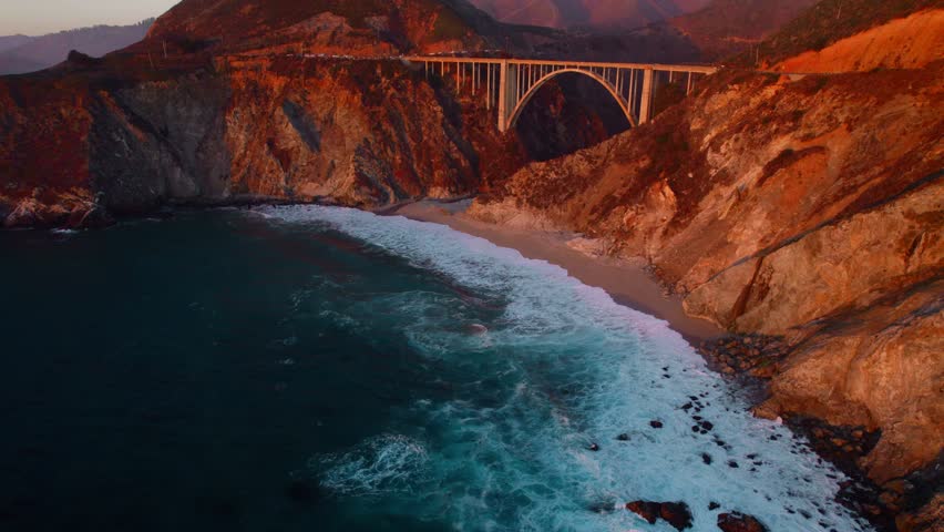 Flying over rocky coastline along cliffside as the light hits the surface during sunset. Bird eye view 4K cinematic drone footage. Soar above sea coast from up in air. Blue ocean waves crashing.