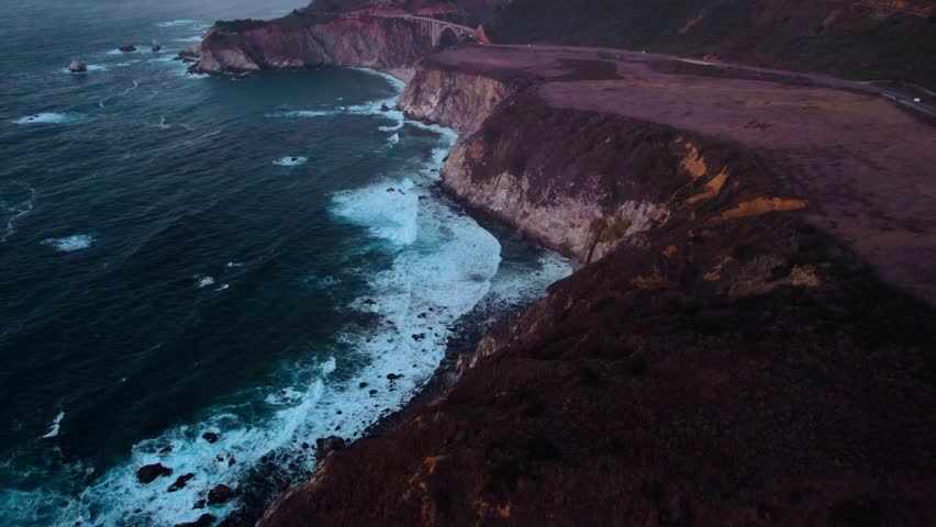 Flying over rocky coastline along cliffside as the light hits the surface during sunset. Bird eye view 4K cinematic drone footage. Soar above sea coast from up in air. Blue ocean waves crashing.