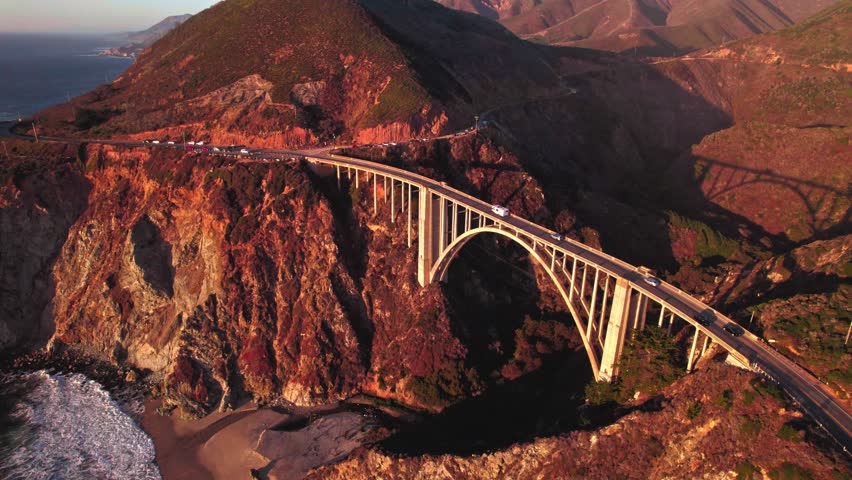 Flying over rocky coastline along cliffside while the sunlight hits the bridge during sunset. Bird eye view 4K cinematic drone footage. Soar above sea coast from up in air. Blue ocean waves crashing.