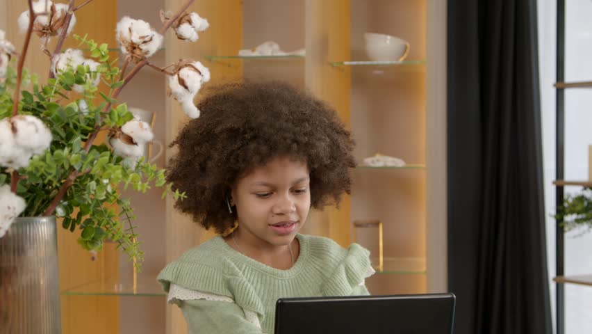 Portrait of joyful positive adorable elementary age African American girl with curly hair in handsfree device talking with friend, networking and browsing funny video content online using tablet pc.