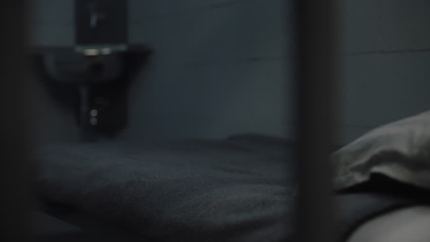 Inmate puts orange prison uniform, bath towel and toilet paper on bed. Guilty criminal or prisoner serves imprisonment term in jail. Detention center or correctional facility. View through metal bars.