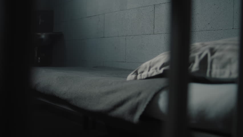 Prisoner puts orange uniform and bathroom accessories on the bed in prison cell. Criminal, inmate serves imprisonment term in jail. Detention center or correctional facility. View through metal bars.