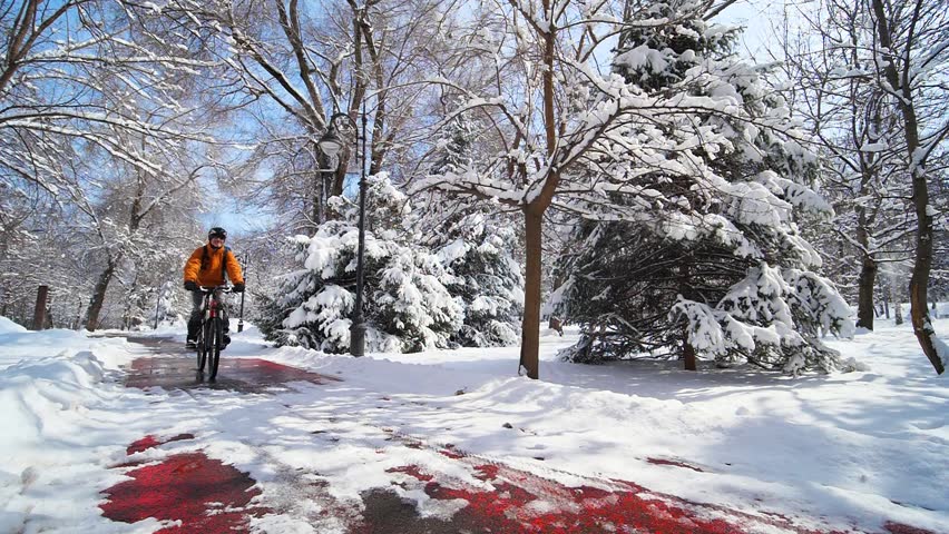 A cyclist rides along a bike path in a city park among snow-covered trees on a sunny winter day. Active lifestyle in winter. Eco-friendly transport. Slow motion 2x