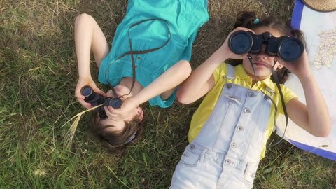 two Young little cute Girls playing explorers Looking at Sky with binoculars lying on grass in green field with map of italy slow motion