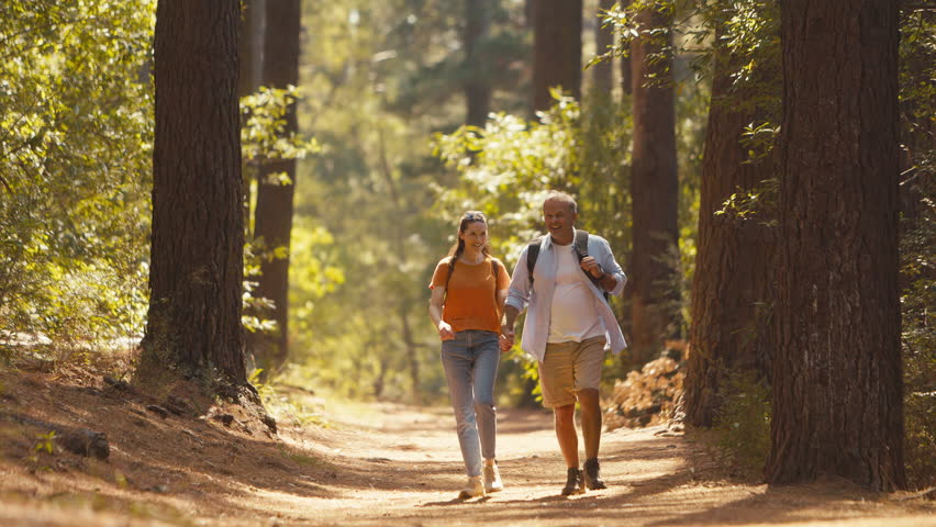 Loving retired senior couple wearing backpacks holding hands hiking along trail through summer countryside - shot in slow motion