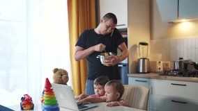 father cooking in kitchen remote work. baby twins playing with laptop in the kitchen. father working at home in the kitchen stirring food in the pan. lifestyle remote work business concept