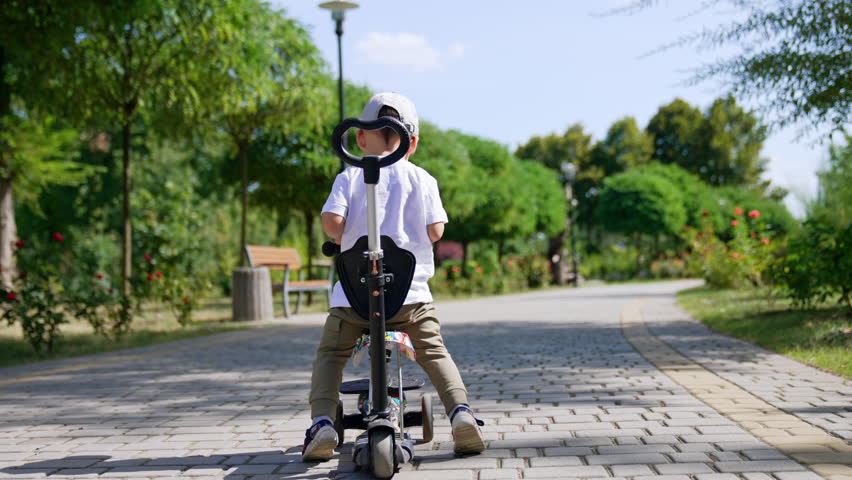 Rear view of a toddler distancing from a camera on his bike. Kid pushes from the ground to move by the paved road in the park.