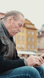 Vertical video, Elderly man in a leather jacket sits on the steps of the monument and looks carefully into a smartphone at Palace Square in Warsaw, Poland, Slow motion