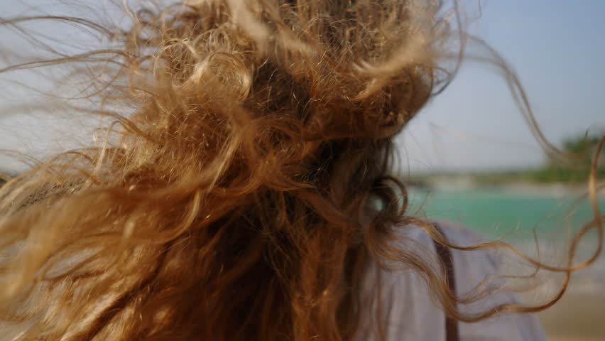 Healthy long hair blowing in wind. Pretty curly model with airy, strong hair posing at coast, sea surf. Young caucasian woman with natural, shiny hair enjoy sunny day at the ocean shore. Close-up.
