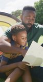 Vertical video of african american father and son sitting in tent and reading, in slow motion. Spending quality time, domestic life and childhood concept.