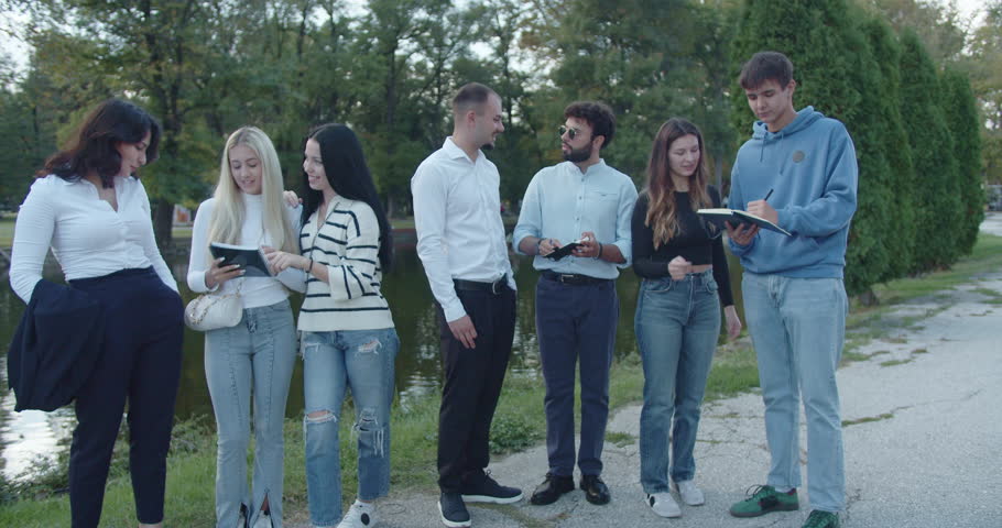 A youthful, mixed-gender group engages with smart phones and tablets outdoors, embodying technology use and connectivity in a natural park environment.