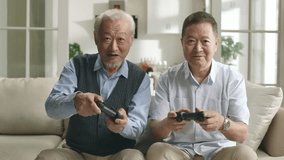 two happy senior asian men sitting on couch at home playing video game together