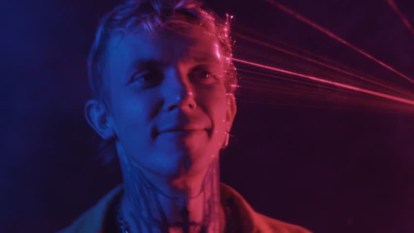 Medium close up of cheerful young man with tattoo on neck and earring partying in nightclub in neon light