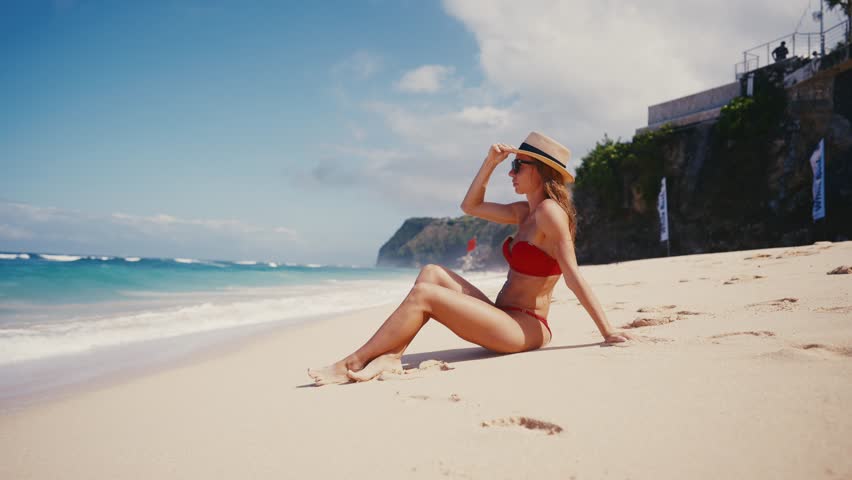 Blonde woman in red bikini straw hat sitting on ocean beach in tropical paradise island with turquoise water waves, white sand in sunny weather. Girl admire seascape. Beach resting, luxury tourism.