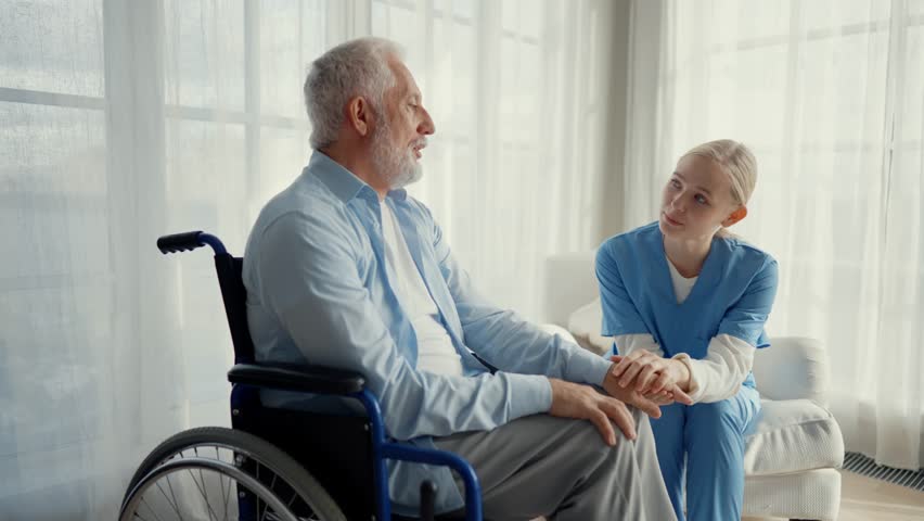 Old man in wheelchair talks with nurse. Senior paralyzed male thanks social worker woman for help support at clinic. Caregiver holds hand patient shoulder stands near. Legs paralysis, special needs. Royalty-Free Stock Footage #3461556451
