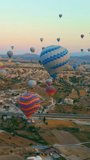 Vertical video. In this aerial video, the skies above Cappadocia, Turkey, come alive with a kaleidoscope of hot air balloons. Against the backdrop of the region's iconic valleys, rocks, and fields