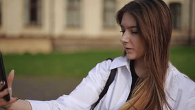 Slow motion. A beautiful girl with long hair takes a selfie on a mobile phone, standing against the background of the building, in which she studies
