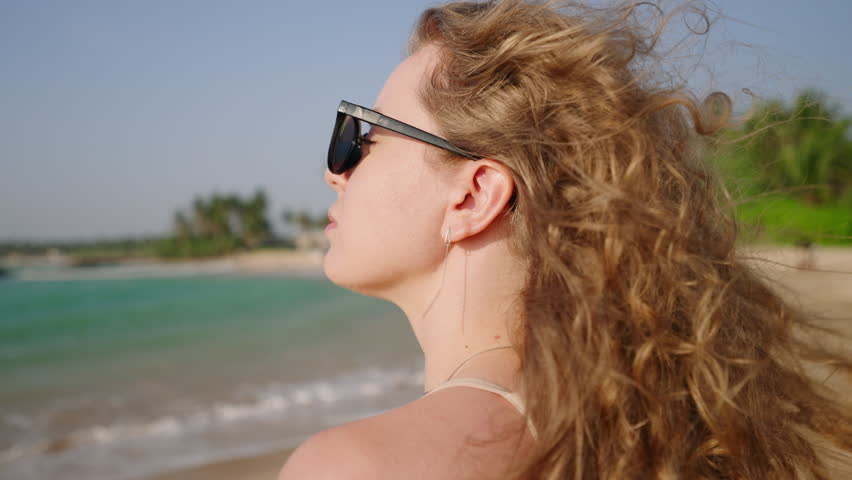 Healthy long hair blowing in wind. Pretty curly model with airy, strong hair posing at coast, sea surf. Young caucasian woman with natural, shiny hair enjoy sunny day at the ocean shore. Close-up.