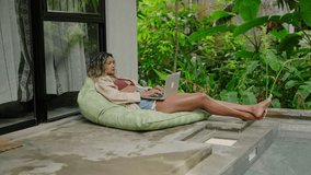Relaxed biracial woman works on laptop, lounges on bean bag by pool in tropical garden. Freelancer enjoys remote job, weekend leisure amidst green plants. Slow motion. - Powered by Shutterstock - Get 15% off with code: PIKWIZARD15