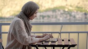 Young muslim woman wearing hijab using technology indoors on balcony. Horizontal panoramic view of arabic woman working with laptop at home. Technology and women lifestyle. Modern multicultural life