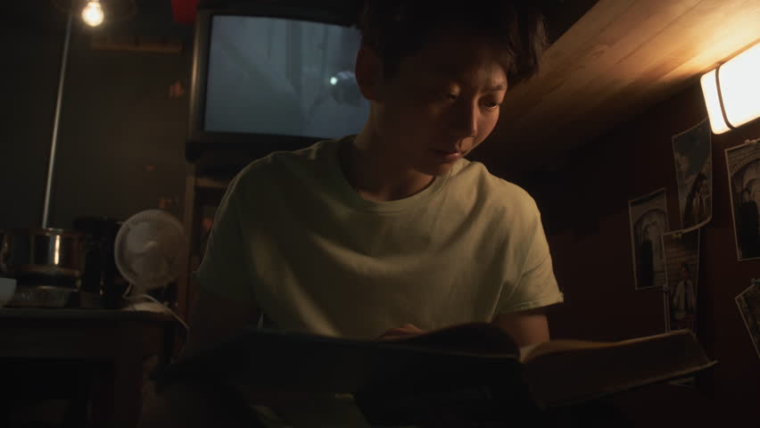 Medium close-up shot of young androgynous Asian woman or man sitting on bunk bed in dimly lit micro flat, reading textbook, revising material for university exam, movie on old TV in background Royalty-Free Stock Footage #3461892063