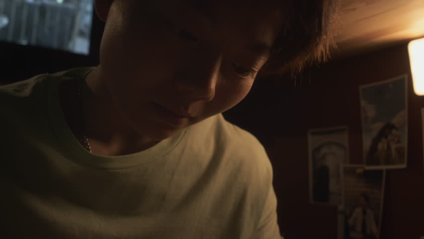 Close-up shot of young androgynous Chinese female student with short hair sitting in bedroom at night, reading textbook, revising and learning material for university exam, movie on TV in background Royalty-Free Stock Footage #3461892079