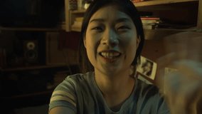 Close-up UGC selfie shot of cheerful Asian female student recording video message for friend or family on smartphone, while sitting in micro dorm flat in evening, to share good news