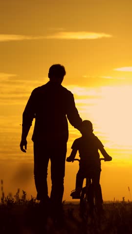 Father helps his kid son ride bike. Father teaching little son to ride bike in park. Child rides bike. Child, dad play together, sunset. Child dream. Kid boy learns to ride bike. Happy family walk sun