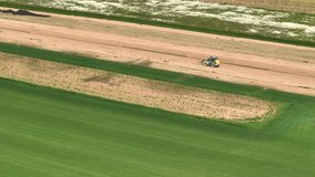 AERIAL: Forklift drives along a patch of parched dry soil running alongside a sod field watered by sprinklers. Forklift driving across a field of turf grass growing at a large industrial sod farm.