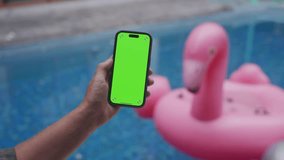 POV male hand holding smartphone with empty green screen chroma key relaxing near pool with pink flamingo background  - Powered by Shutterstock - Get 15% off with code: PIKWIZARD15