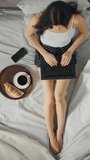 Young Millennial Girl Sitting on a Bed in the Morning, Uses Laptop Computer and Eats Croissants, Drinks Coffee for Breakfast. Top Down Zoom Out. Video Footage with Vertical Screen Orientation 9:16