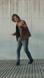 Cheerful and Happy Young Man with Long Hair Actively Dancing While Walking Down the Stairs. He's Wearing a Brown Leather Jacket. Vertical Screen Orientation Video 9:16