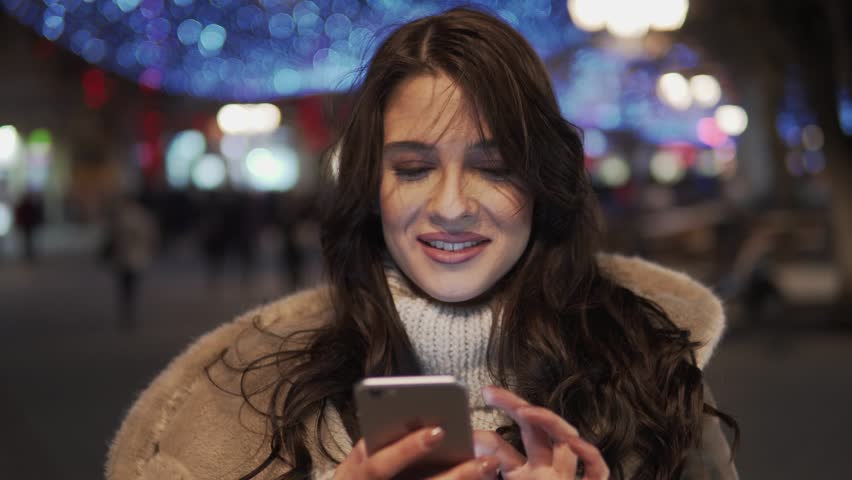 Beautiful young woman using smart phone and smiling. Typing message, looking at mobile and smiling. Using mobile phone outside at the city center.